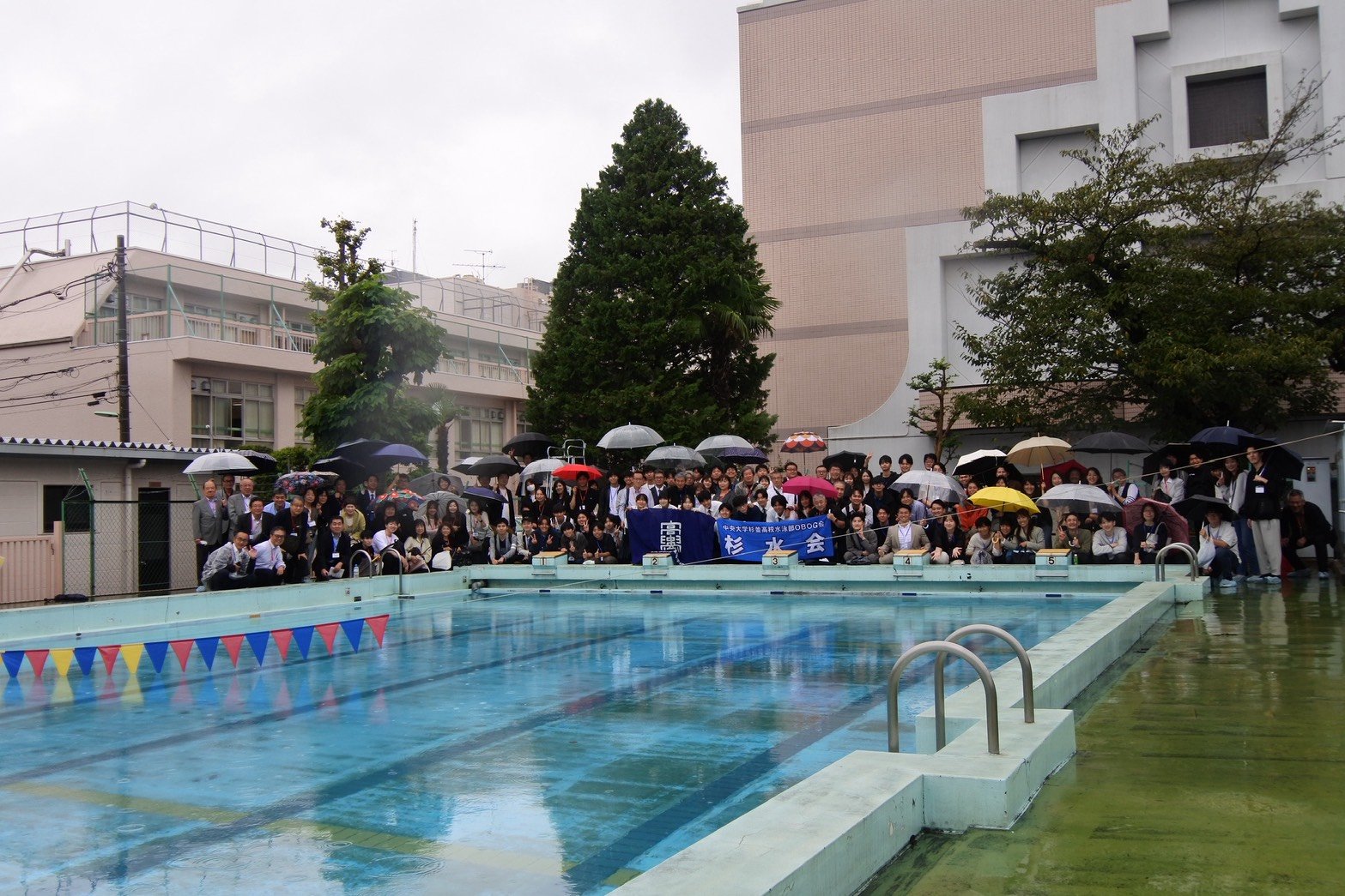 雨のプールサイドで、杉水会の旗を囲む参加者の記念撮影